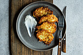 Overhead view of crispy golden potato latkes served with a dollop of sour cream on a rustic ceramic plate, accompanied by cutlery.