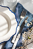 An elegant table setting featuring a white plate with a blue-rimmed edge, silver cutlery, and delicate cherry blossoms on a textured tablecloth