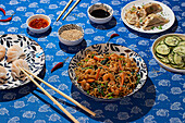 A vibrant assortment of Chinese food served on decorative dishes. Featured are noodle dish with shrimp, steamed dumplings, cucumber salad, and sauces