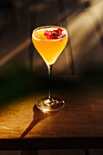 A sophisticated cocktail adorned with a vibrant red flower garnish, cast in a warm golden light on a wooden surface, projecting an elegant shadow