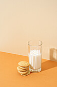 Ein ordentlicher Stapel knuspriger Haferflockenkekse neben einem vollen Glas Milch auf einer orangefarbenen Fläche, mit weichen Schatten.