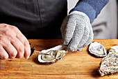 A chef's hands, wearing protective gloves, expertly opening fresh oysters with a specialized knife on a wooden board.