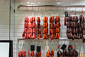 A variety of chorizos and sausages are displayed hanging on metal hooks against a tiled wall in a deli