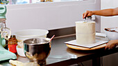 A person meticulously applies frosting to a tall cake, with baking equipment and ingredients dispersed around the kitchen workspace