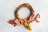 A rustic autumn wreath made of intertwined branches, adorned with scattered dried leaves in shades of red, yellow, and brown, presented on a gray textured background