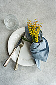 Elegant dining setup featuring a white ornate plate, vintage silverware, a blue linen napkin, and a bright yellow mimosa bouquet on a textured backdrop