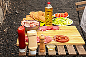 A ready-to-grill outdoor barbecue selection featuring raw meats, buns, and salads on a wooden board, complete with condiments and a bottle of oil.