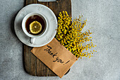 A top view of a classic tea cup with lemon, placed on a wooden board, accompanied by bright Mimosa flowers and a Thank You! card on a textured background