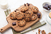 Freshly baked chocolate chip cookies on a wooden cutting board, accompanied by dark chocolate pieces and a glass of milk