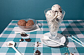 Elegant glass filled with cookies and cream ice cream, accompanied by a bowl of chocolate scoops, set on a blue checkered tablecloth with scattered cookie pieces and candy sprinkles