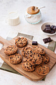 Freshly baked chocolate chip cookies arranged neatly on a wooden cutting board, accompanied by a jug of milk and sugar contained in a jar with a wooden lid, all set on a light textured surface