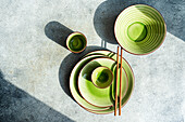 Top view of a stylish table setting featuring green ceramic dishes, bowls, and a cup with brown chopsticks, all bathed in natural sunlight with shadows on a textured grey surface.