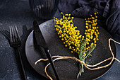 From above a sophisticated black plate setting accented by a fork, knife, and a bright spray of yellow mimosa flowers bundled with twine.