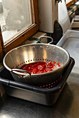 Fresh strained tomatoes sit in a stainless steel colander within a plastic container by a kitchen window, capturing steps in pizza sauce preparation