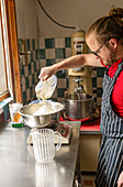 A skilled chef, wearing a striped apron, prepares pizza dough by pouring flour into a metal mixing bowl in a kitchen equipped with vintage machinery