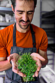 A man, wearing an orange shirt and black striped apron, carefully holds a lush container of vibrant microgreens, smiling with satisfaction at his indoor garden