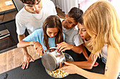 Three triplets pour popcorn into a bowl in a modern kitchen, engaging in teamwork and enjoying a shared activity