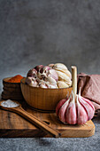 A carefully arranged set of cooking ingredients featuring fresh garlic bulbs in a wooden bowl, accompanied by spices and a salt spoon, set against a textured gray backdrop