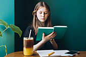 Female student sitting at a table, absorbed in reading a green book, with iced tea and a smartphone nearby. She is looking down, engaged in her study in a calm setting.