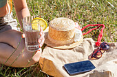 Nahaufnahme einer Hand, die ein Glas Limonade mit einer Zitronenscheibe hält, ergänzt durch sommerliche Utensilien wie einen Strohhut, eine Sonnenbrille und ein Mobiltelefon, alles auf einer Graswiese