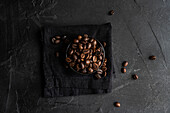 Top view of dark roasted coffee beans elegantly arranged in a small metal bowl atop a textured black napkin and dark grey, rough surface. The scattered beans enhance the visual appeal, emphasizing quality and freshness.