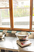 A digital kitchen scale with a bowl of flour on a stainless steel countertop, ready for pizza dough preparation, with a bright window view