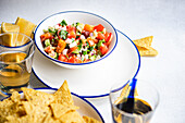 Bunte und würzige mexikanische Salsa mit Tomatenwürfeln, Zwiebeln, Koriander, Avocadostücken und einem Spritzer Zitronensaft in einer weißen Schale mit blauem Rand, dazu Mais-Chips und ein Glas Weißwein