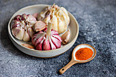 A close-up of fresh garlic bulbs and cloves displayed in a ceramic dish alongside a wooden spoon of vibrant spices, set against a textured gray background