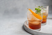 Elegant cherry liqueur cocktails mixed with tonic, accented with ice, a fresh orange slice, and mint leaves, set against a soft grey backdrop