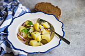 Traditional German potato salad featuring boiled potatoes mixed with slices of red onion and fermented cucumber, presented in a decorative white and blue bowl, accompanied by slices of rye bread