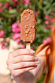 Nahaufnahme einer Hand, die ein schokoladenüberzogenes Mandeleis hält, hervorgehoben vor einem leuchtenden Hintergrund aus rosa Blumen, die die Freude und Frische des Sommers symbolisieren