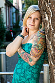 Portrait Young Woman with Tatoos in leaning aginst Tree in Residential Garden. Tilburg, Netherlands.