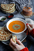 Woman's hands holding a bowl of soup and a piece of pita bread, with spices and Turkish tarhana in a glass.