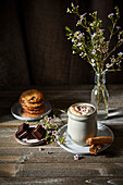 Mexican hot chocolate with whipped cream on a dark wooden background with flowers, biscuits and cinnamon sticks.