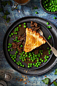 A slice of venison meat pie on a plate served with peas and gravy.