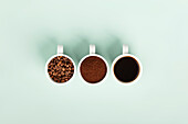 Three white cups with coffee beans, coffee grounds and brewed coffee arranged on a mint-coloured surface