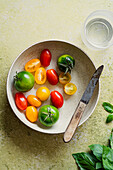 Bowl of small, colourful char and plum tomatoes with a small knife on a green background