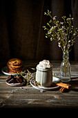 Mexican hot chocolate topped with whipped cream, on a dark wooden background with flowers, biscuits and cinnamon sticks.