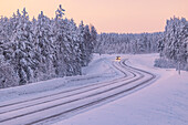 Ein Auto fährt auf einer schneebedeckten Straße durch den Wald in Finnisch-Lappland während einer blauen Stunde im Winter, Muonio, Finnland, Europa