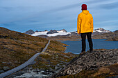 Mann in gelbem Regenmantel mit Blick auf Bergpass, Gletscherberge und Schmelzwasserseen, Jotunheimen Nationalpark, Norwegen, Skandinavien, Europa