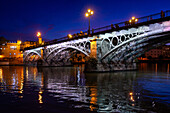 Die bei Einbruch der Dunkelheit beleuchtete Triana-Brücke mit Spiegelungen auf dem Fluss Guadalquivir in Sevilla, Andalusien, Spanien.