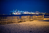 Holzzaun am Strand von El Rinconcillo mit dem beleuchteten Hafen von Algeciras im Hintergrund während der Abendzeit.