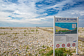 Weitwinkelansicht des felsigen Strandes Plage Du Havre in Le Havre, Frankreich, mit einem klaren Schild mit Strandhinweisen und einem teilweise bewölkten Himmel.