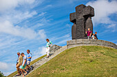 Eine Gruppe von Besuchern erkundet den deutschen Soldatenfriedhof in der Normandie, Frankreich, mit einem großen Kreuzdenkmal im Hintergrund.