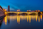 Atemberaubende Nachtansicht der beleuchteten Triana-Brücke, die sich im ruhigen Wasser des Guadalquivir-Flusses in Sevilla, Spanien, spiegelt.