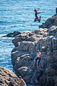 A young individual jumps off a rocky cliff into the sea in Ouistreham, Brittany, France, conveying a sense of adventure and excitement.