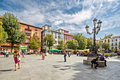 Belebte Szene auf der Plaza de Bib Rambla, Granada, Spanien, mit Menschen, die einen sonnigen Tag umgeben von historischen Gebäuden genießen.