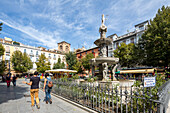 View of tourists walking and enjoying the sunny day at Plaza de Bib Rambla in Granada, España with a beautiful fountain and historic buildings.