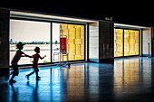 Zwei Kinder spielen und rennen im Terminal des Aeropuerto de Sevilla, Spanien. Das Sonnenlicht erhellt den Innenraum und schafft eine fröhliche und lebhafte Atmosphäre.