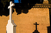 Weißes Kreuz wirft einen Schatten auf eine Backsteinmauer am Cementerio de San Fernando in Sevilla, Andalusien, bei Sonnenuntergang.